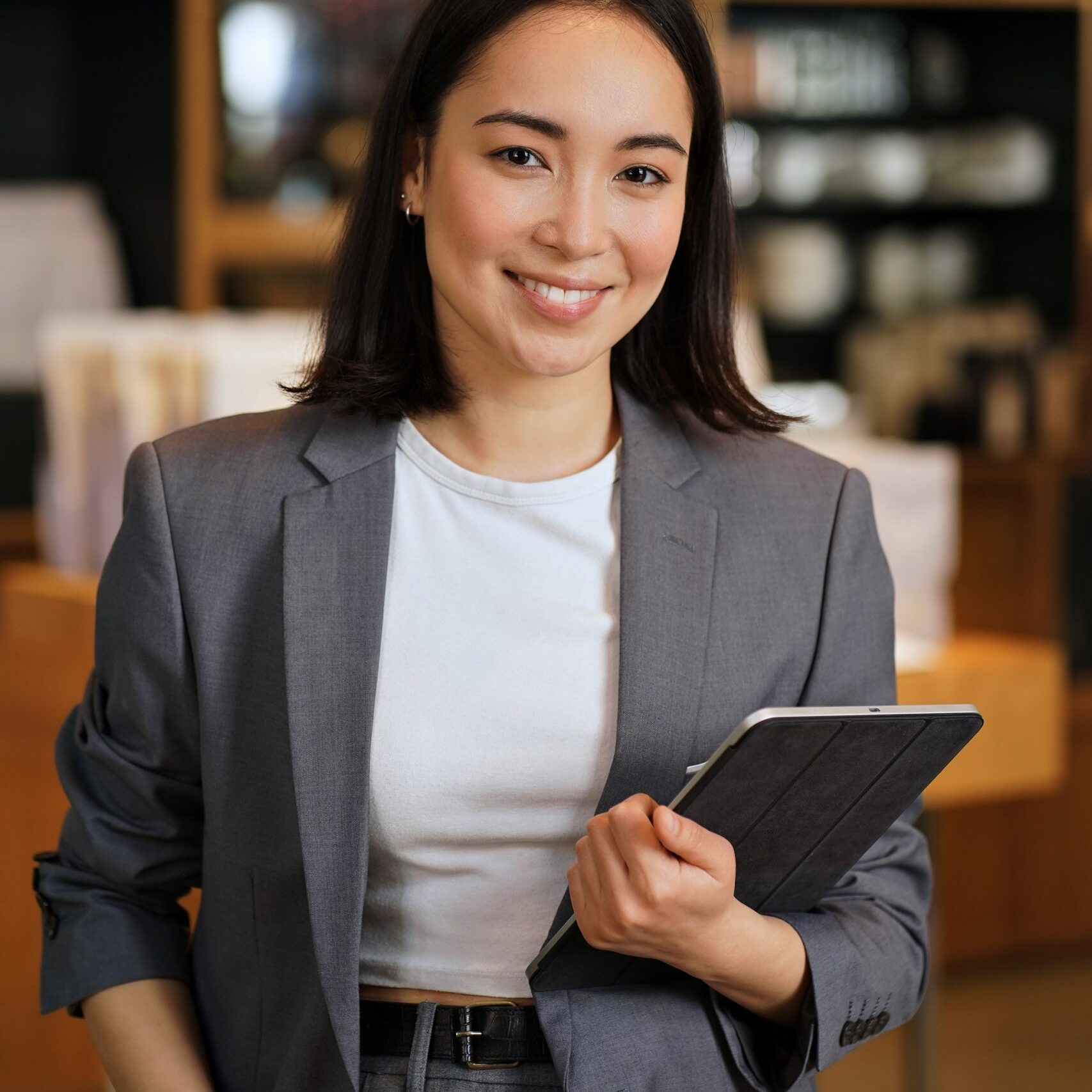Young smiling successful professional leader Asian business woman, female executive manager, saleswoman wearing suit holding digital tablet standing in office looking at camera, vertical portrait.